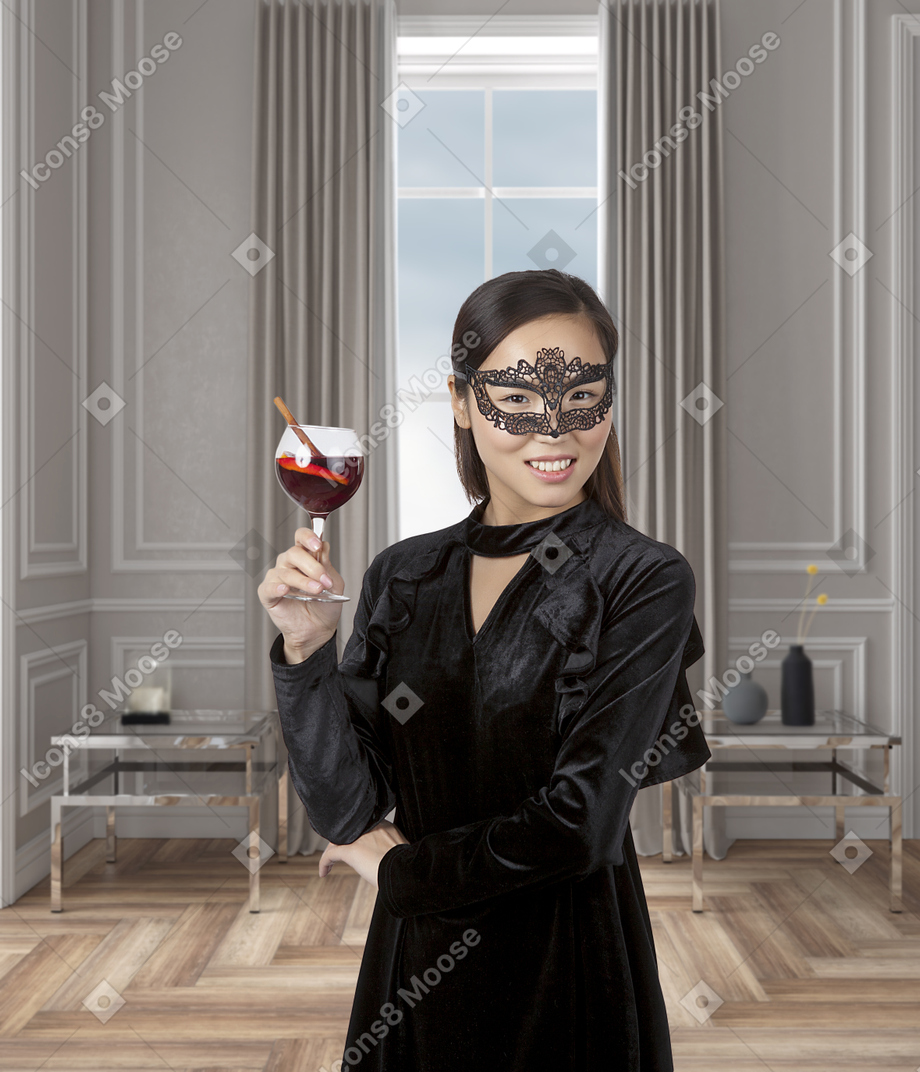 美丽的亚洲女人在黑色天鹅绒连衣裙和化妆舞会面具站在一个现代风格的房间，一杯鸡尾酒在她的手