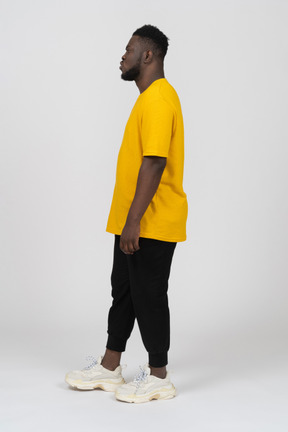 Vista laterale di un giovane uomo imbronciato dalla pelle scura con una maglietta gialla in piedi immobile