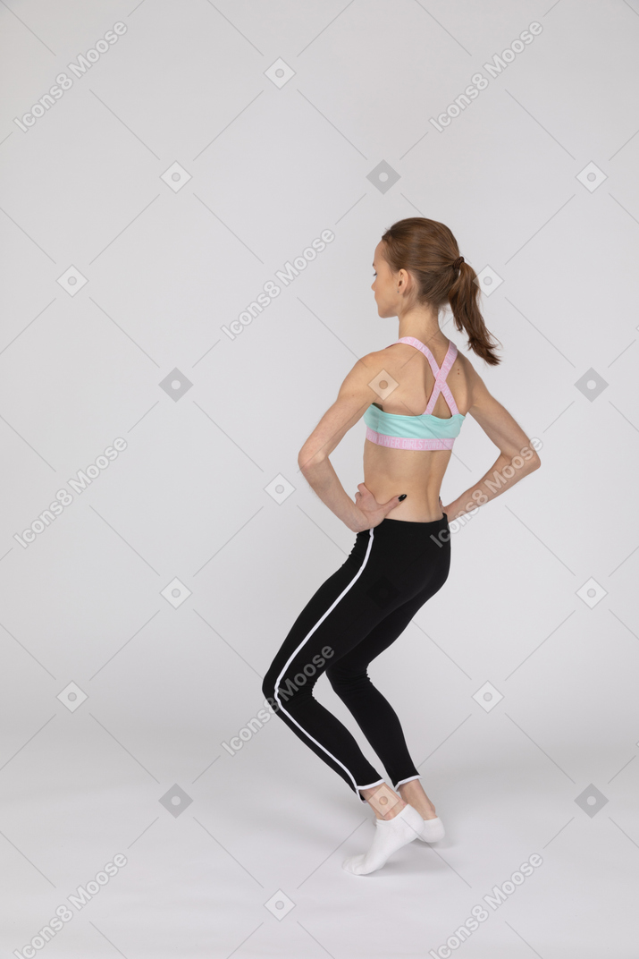 Vista posterior de una jovencita en ropa deportiva poniendo las manos oh caderas y doblando las rodillas