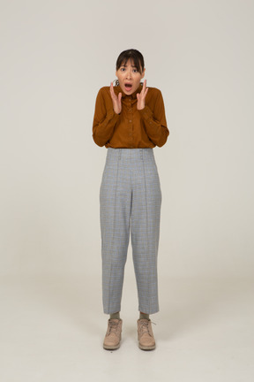 Front view of a shocked young asian female in breeches and blouse raising hands
