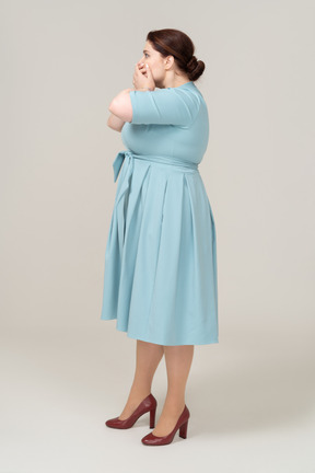 Вид сбоку испуганной женщины в синем платье, закрывающей рот руками