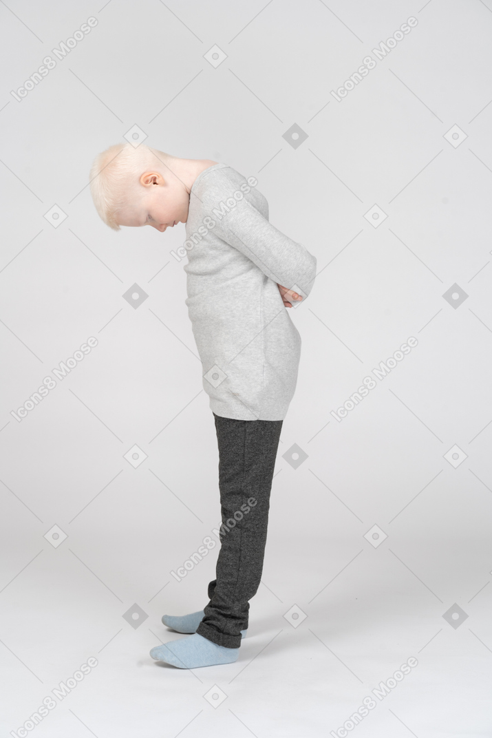 A blonde little boy in casual clothes standing and hiding hands behind nodding head