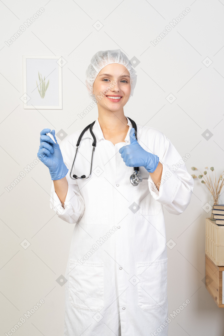 Front view of a smiling young female doctor with stethoscope holding thermometer and showing thumb up