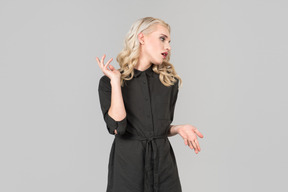 A young blond-haired person in a black dress  standing against the plain grey background