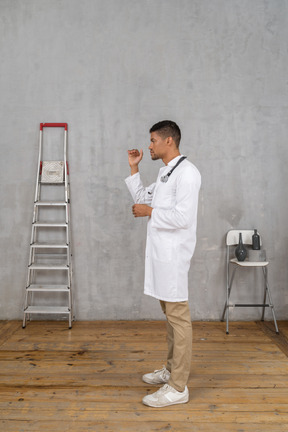 Vista lateral de un joven médico de pie en una habitación con escalera y silla mostrando el tamaño de algo