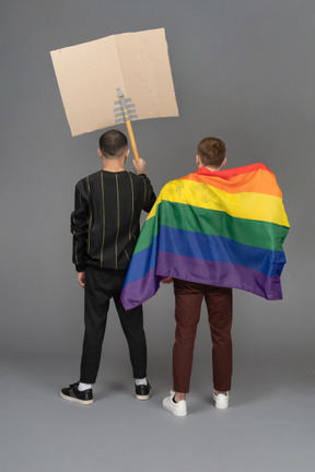 Vista posteriore di due giovani uomini in possesso di un cartellone pubblicitario e indossando la bandiera lgbt