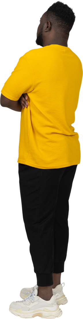 Vista posterior de tres cuartos de un joven de piel oscura con camiseta amarilla cruzando los brazos