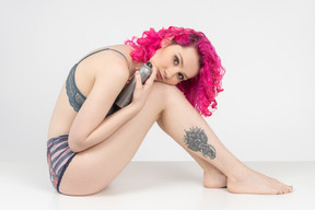 Linda garota de cabelo rosa com perna tatuada sentada no chão com uma garrafa de prata