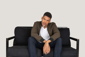 Vorderansicht eines jungen mannes, der auf einem sofa sitzt und eine zigarette im mund hält