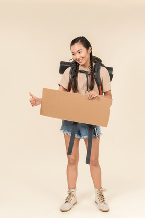 Young female hitchhiker holding paper card and showing thumb up