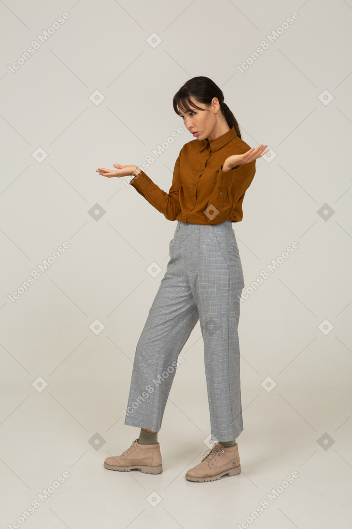 Vista de três quartos de uma jovem mulher asiática questionadora de calça e blusa, levantando as mãos