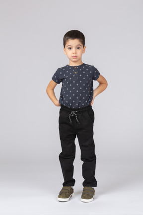 Vue de face d'un garçon mignon dans des vêtements décontractés posant avec les mains sur le dos et regardant la caméra