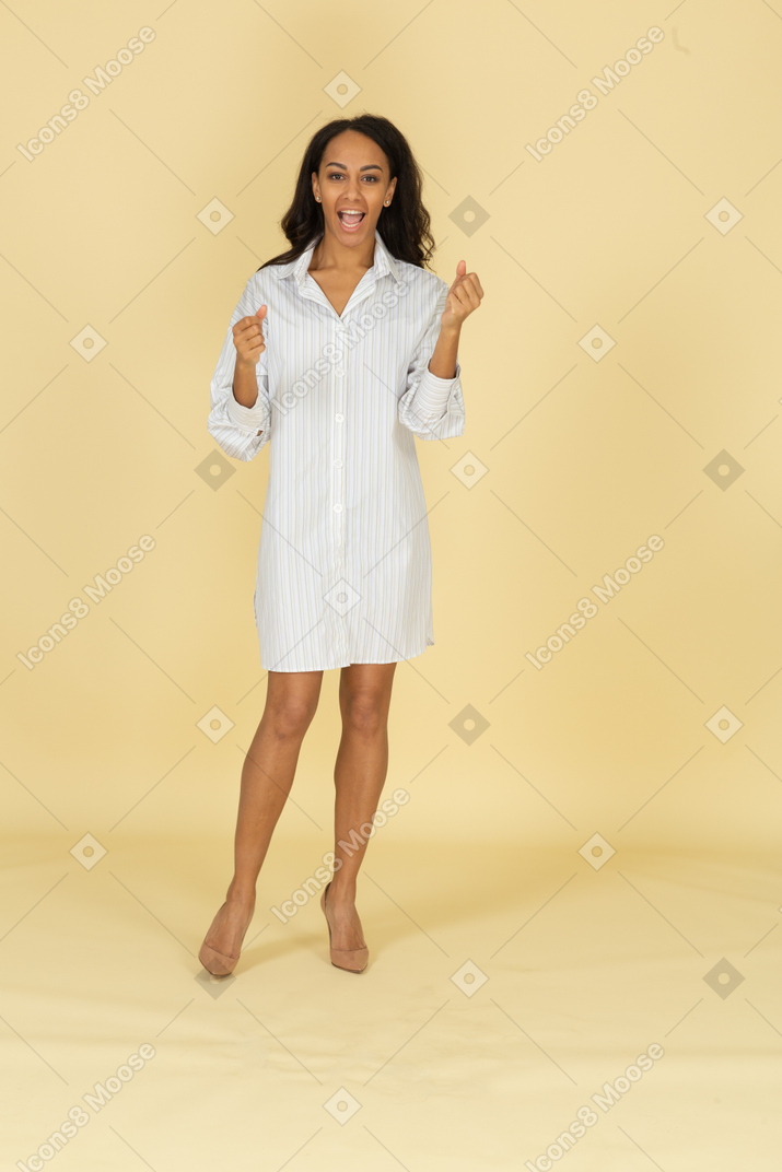 Vista frontal de una mujer joven de piel oscura gritando en vestido blanco apretando los puños