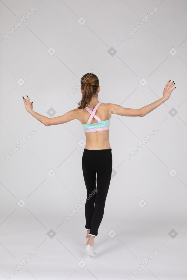 Three-quarter back view of a teen girl in sportswear balancing on tiptoes while raising hands