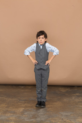 Vista frontal de um menino bonito de terno cinza posando com as mãos nos quadris