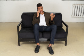 Front view of a perplexed young man sitting on a sofa and talking on his phone