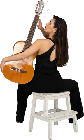 Back view of a sitting young lady in black suit touching the guitar's headstock