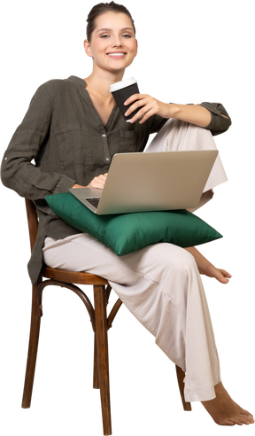 Front view of a smiling young woman sitting on a chair and holding her laptop & coffee cup