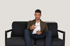 Front view of young man sitting on a sofa and passing pensil with notebook