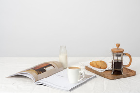 Café en prensa francesa y galletas en la bandeja de madera, botella de leche y taza de café con leche en la revista.