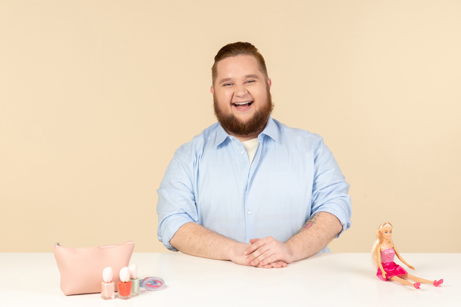Laughing big man sitting at the table with cosmetics and barbie doll on it