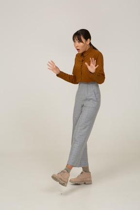 Vista de três quartos de uma jovem mulher asiática cuidadosa de calça e blusa, estendendo os braços
