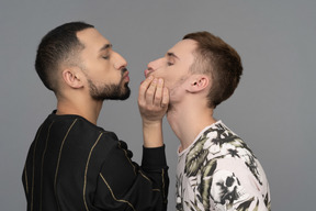 Close-up de um jovem segurando outro homem pela mandíbula, não muito gentilmente como se estivesse tentando beijar