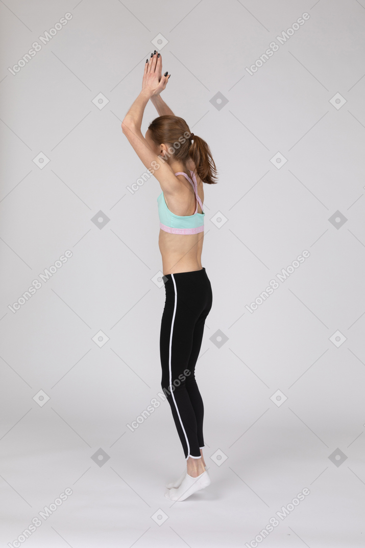 Three-quarter back view of a teen girl in sportswear standing on tiptoes and raising hands