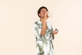 Joven mujer negra de pelo corto en bata, bebiendo de una botella reutilizable