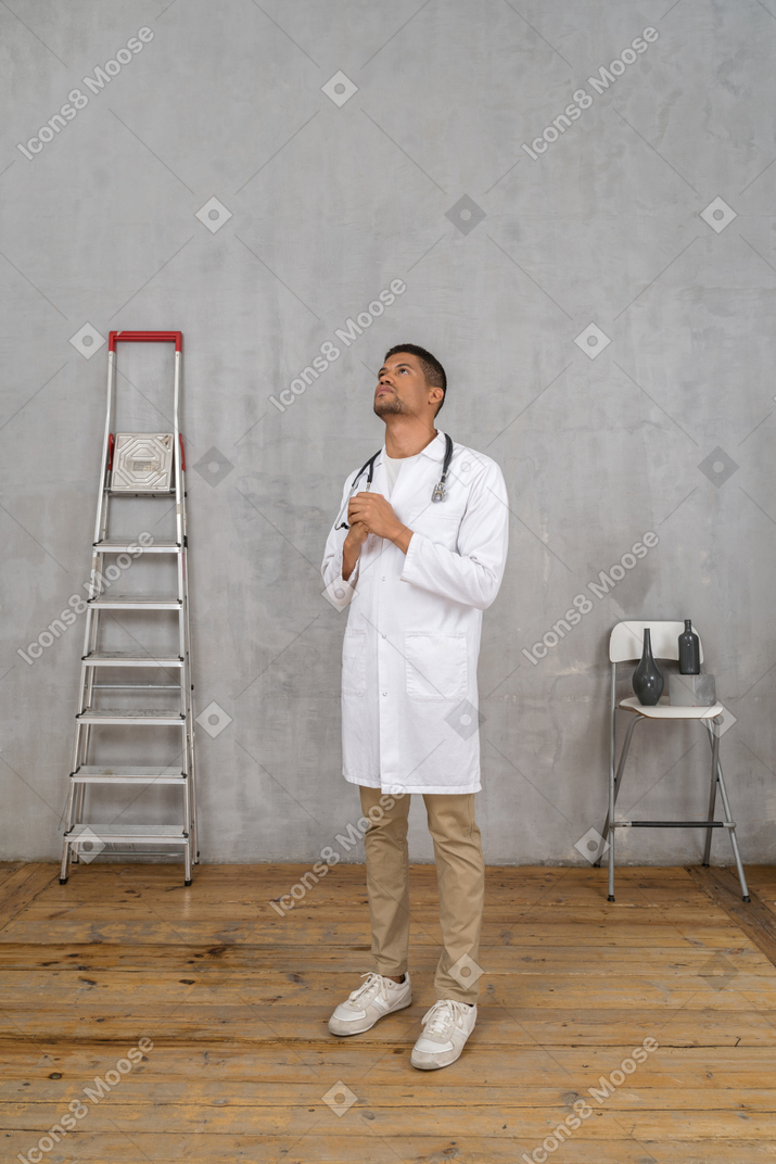 Vista de tres cuartos de un joven médico rezando de pie en una habitación con escalera y silla