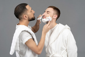 Young caucasian man carefully shaving his partner and putting shaving foam on his nose