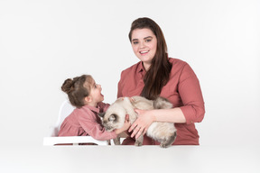 Mother and her little daughter, wearing red and pink clothes, sitting at the dinner table with their family cat