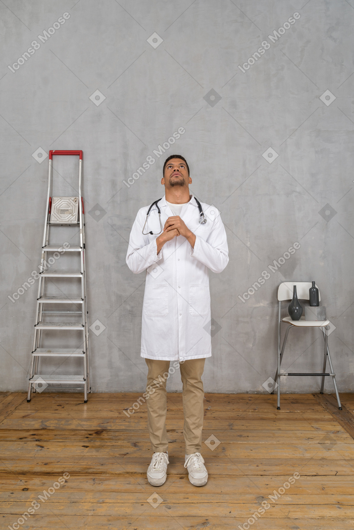 Vista frontal de un joven médico rezando de pie en una habitación con escalera y silla