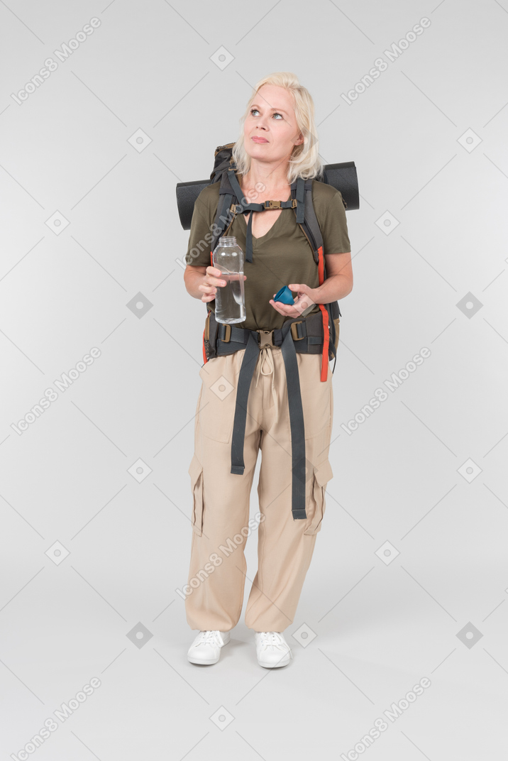 Dreamy mature female tourist carrying backpack and holding bottle of water