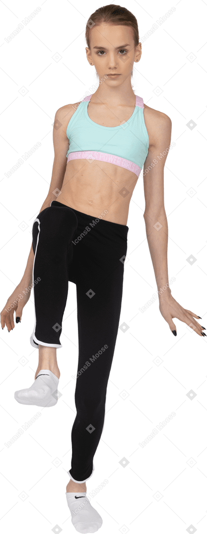 Vista frontal de uma adolescente em roupas esportivas estendendo as mãos e levantando a perna
