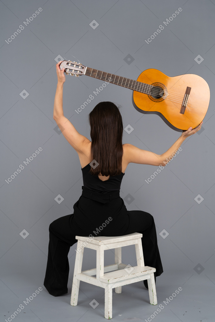 Vista de trás de uma jovem de terno preto segurando o violão e sentada no banquinho