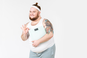 Tipo grande feliz en ropa deportiva mostrando signo bien y sosteniendo pesas digitales
