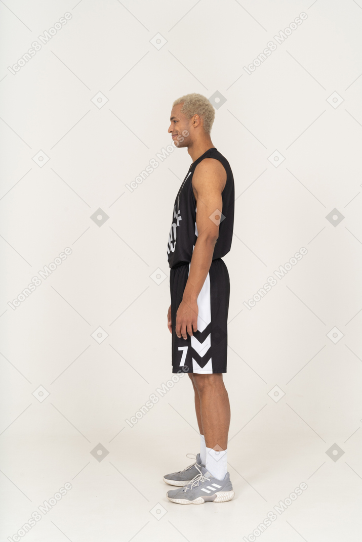 Vista laterale di un giovane giocatore di basket maschile sorridente in piedi immobile