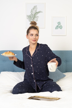 Vista frontal de una joven en pijama sosteniendo una taza de café y algunos pasteles sentado en la cama
