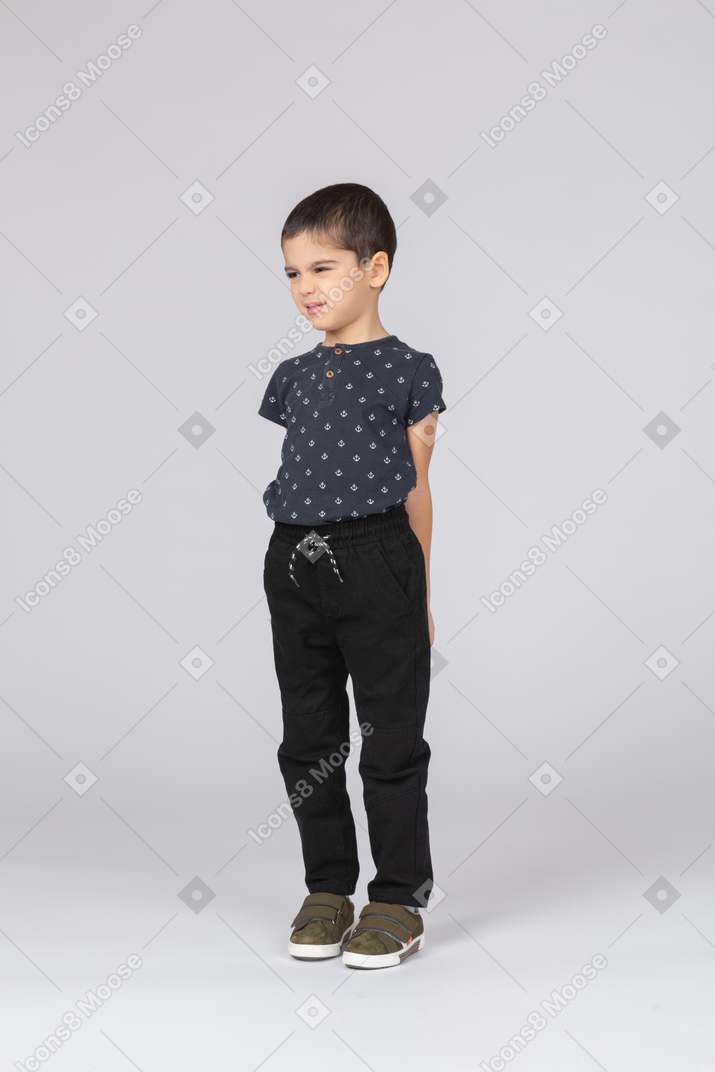Front view of a cute boy in casual clothes standing with hands behind back and making faces