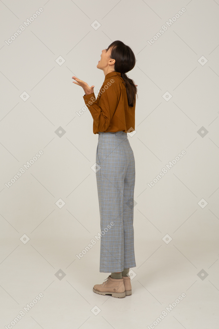 Три четверти сзади удивленной молодой азиатской женщины в бриджах и блузке, поднимающей руки
