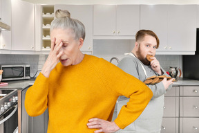 An old woman facepalming and a fat man eating cookies behind her