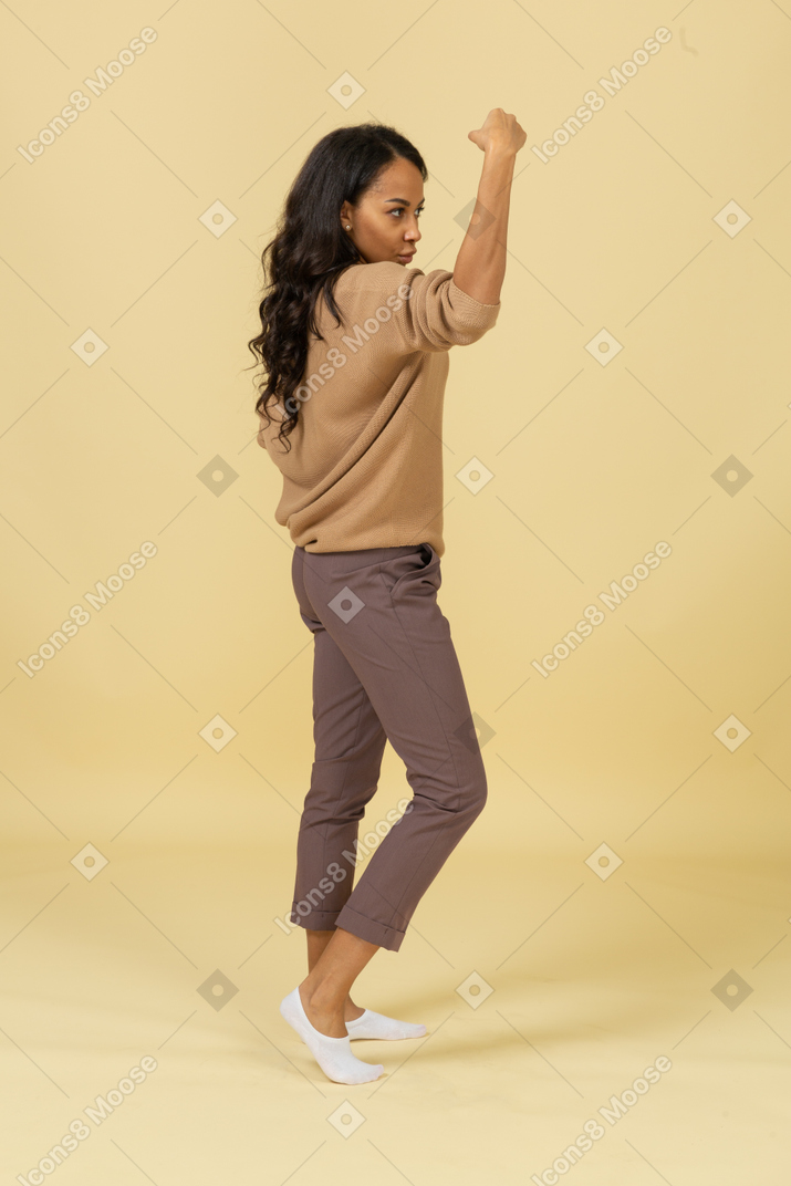Vista lateral de una mujer joven de piel oscura fuerte levantando la mano mientras pone la mano en la cadera
