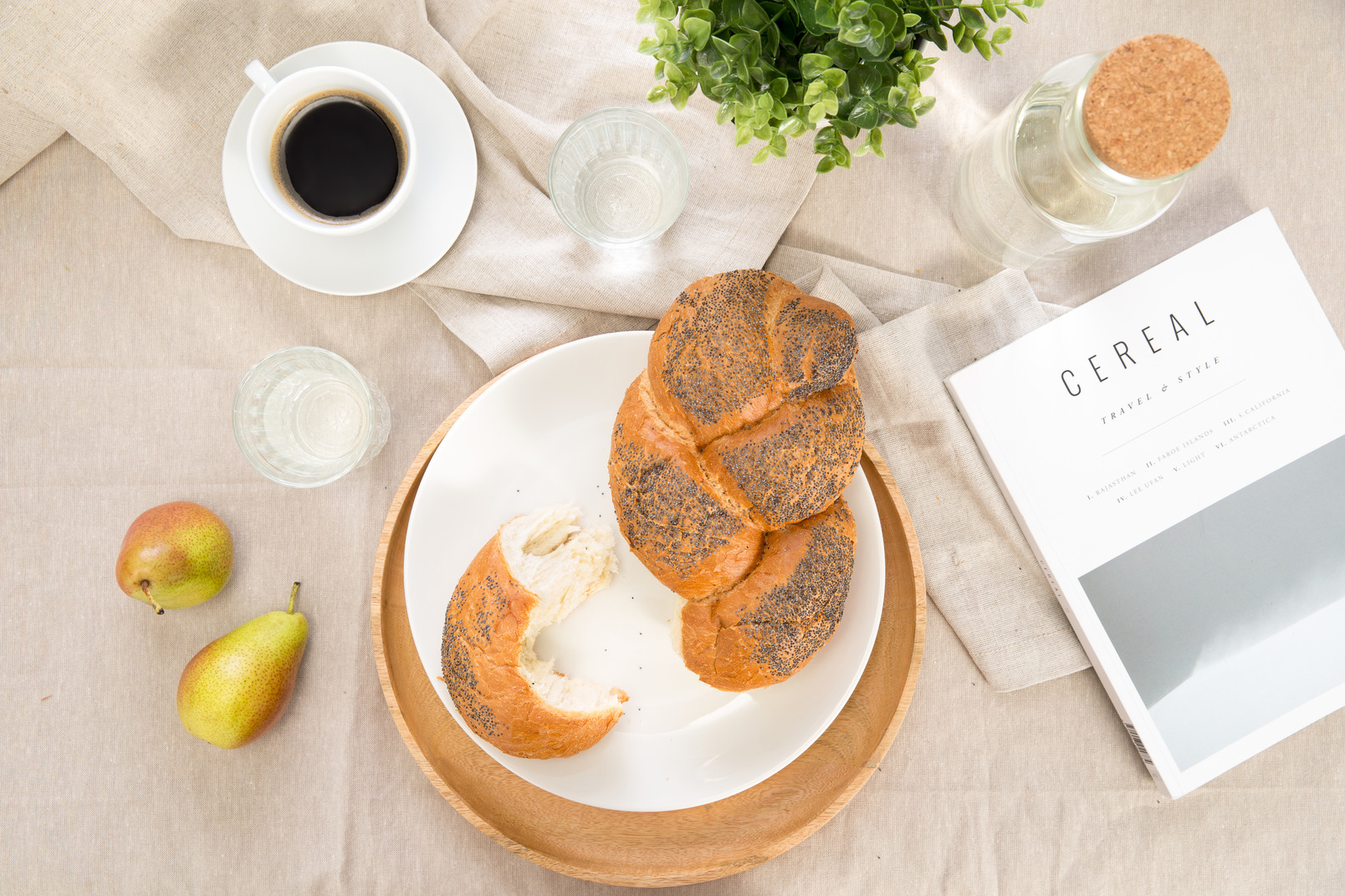 Bread with poppy seeds on the plate, cup of coffee, magazine and water on the tablecloth