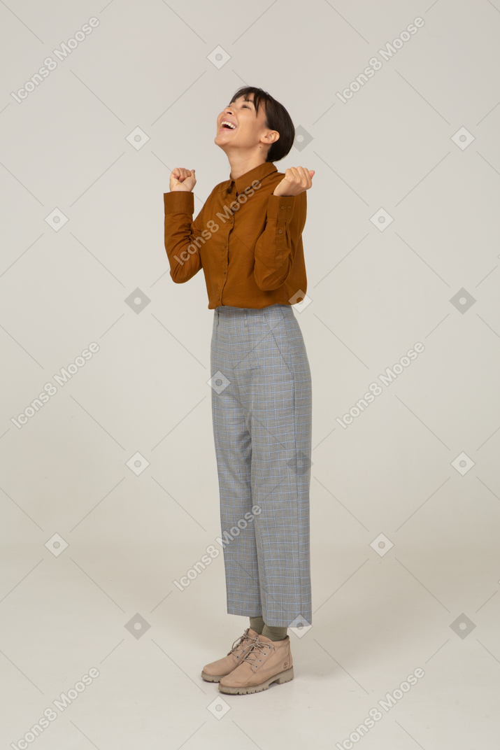 Vista de três quartos de uma jovem asiática encantada de calça e blusa, levantando as mãos