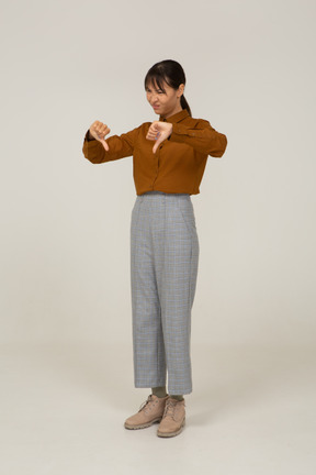 Vista de três quartos de uma jovem mulher asiática de calça e blusa com os polegares para baixo