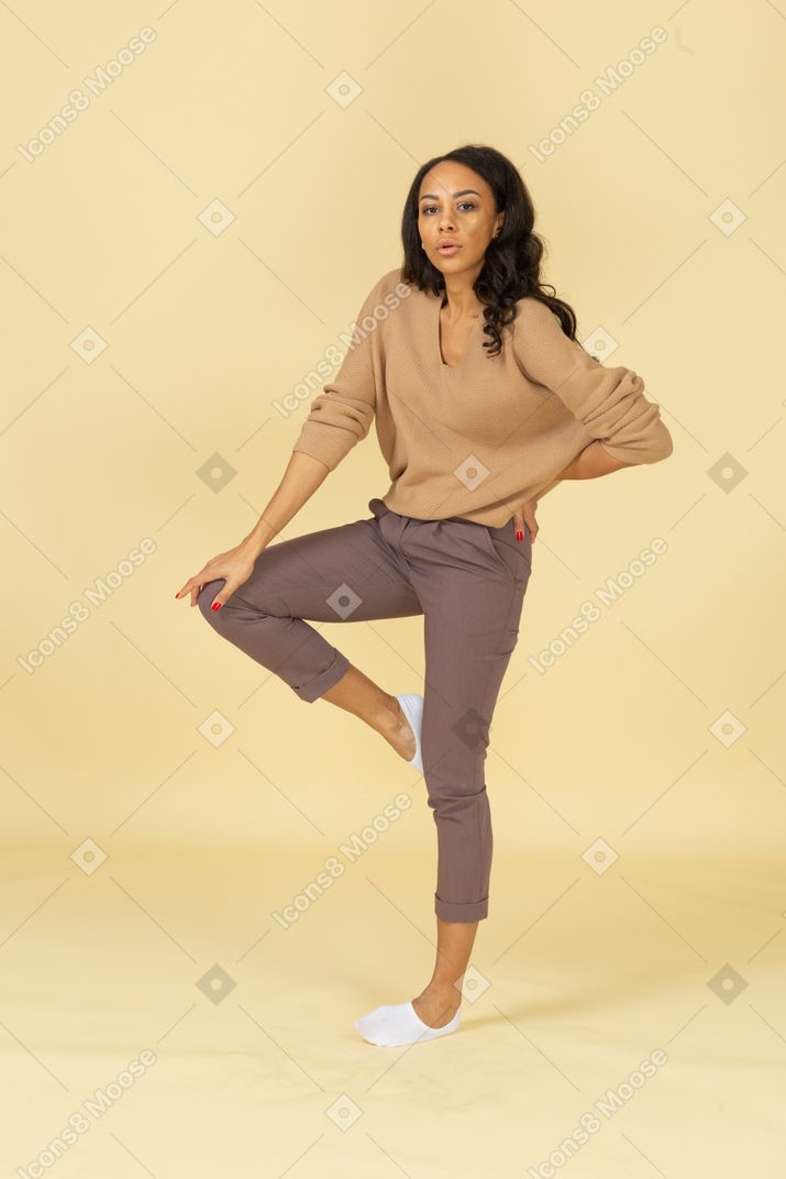 Vista de tres cuartos de una mujer joven de piel oscura levantando la pierna mientras pone la mano en la cadera