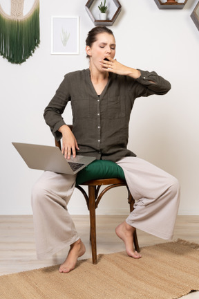Vorderansicht einer müden jungen frau in hauskleidung, die mit einem laptop auf einem stuhl sitzt