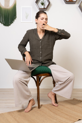 Front view of a tired young woman wearing home clothes sitting on a chair with a laptop