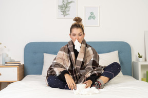 Front view of an ill young lady in pajamas wrapped in checked blanket in bed blowing her nose